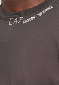 Grå t-skjorte i stoff med ribbestrikket rund hals, dekorert med hvite broderte logoer fra EA7 og Emporio Armani på kragen.