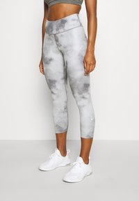 Vrouw draagt grijze tie-dye cropped leggings en witte sneakers, staand tegen een effen witte achtergrond.