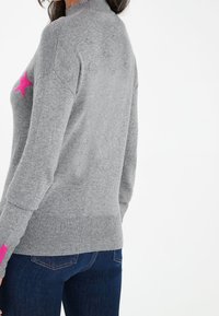 Pull gris avec un motif d'étoile rose sur le devant, présentant un col montant et des poignets côtelés. Porté avec un jean bleu.