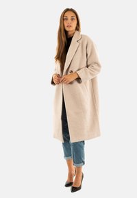 Abrigo de mezcla de lana de color beige claro con solapa con muesca, cierre de un solo botón y bolsillos laterales; combinado con jeans de mezclilla arremangados y tacones negros.