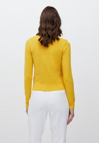 Geel gebreid trui met een ronde hals en ribbelsteek onderaan. De achterkant heeft een gladde textuur en aansluitende mouwen, in combinatie met witte broek.