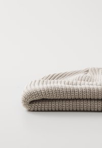 Beige gebreide muts liggen gevouwen. Heeft een geribbelde textuur met een zachte, dikke brei. Eenvoudig ontwerp, neutrale kleur en geen zichtbare patronen of accenten.