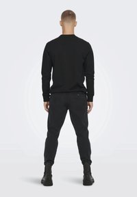 Maglietta nera a maniche lunghe, design aderente, abbinata a pantaloni scuri a gamba affusolata e stivali neri alla caviglia. Texture liscia, silhouette semplice, vista posteriore.