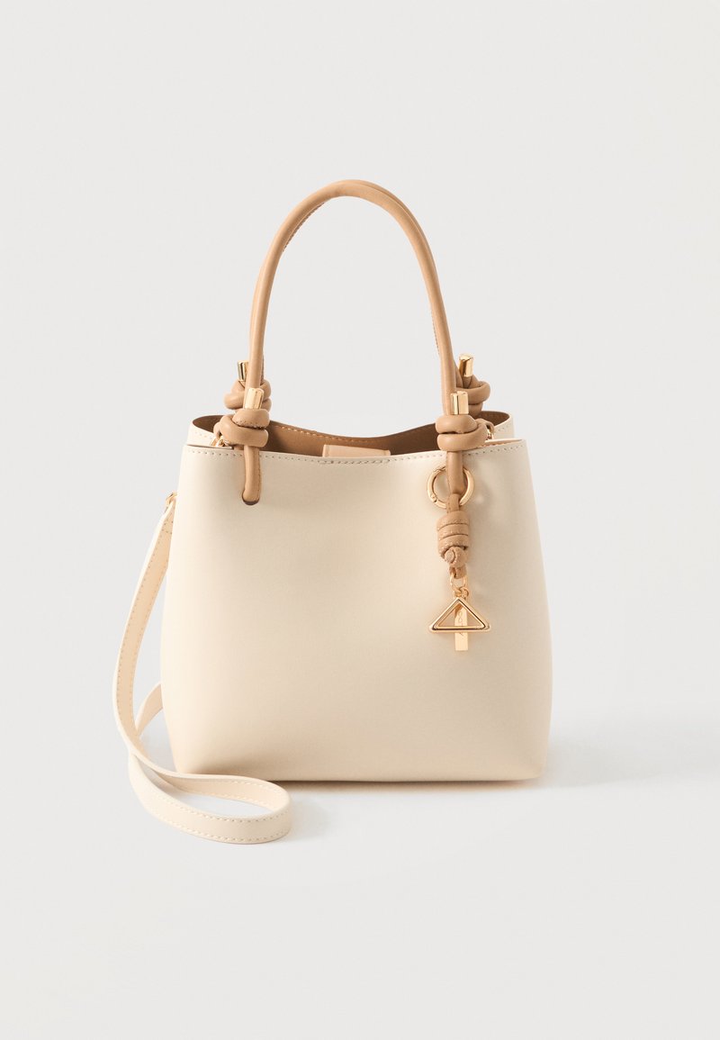 Sac à main couleur crème avec des poignées courtes nouées, une longue bandoulière et un petit charm triangulaire doré suspendu à une poignée.