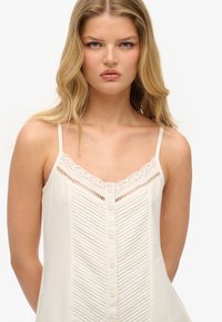 Lichtbeige mouwloze top met kanten afwerking, verticale textuurdetails en een rij knopen aan de voorkant. Zachte stof en relaxte pasvorm.