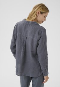 Chemise en lin gris avec des manches longues, détail de empiècement au dos, poignets boutonnés et coupe décontractée. Texture douce et léger éclat.