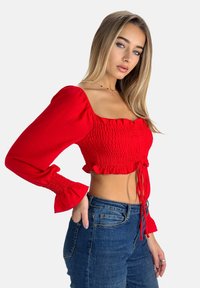 Blusa roja corta con textura fruncida, amplias mangas abullonadas y dobladillo con volantes. Combinada con jeans de mezclilla azul.