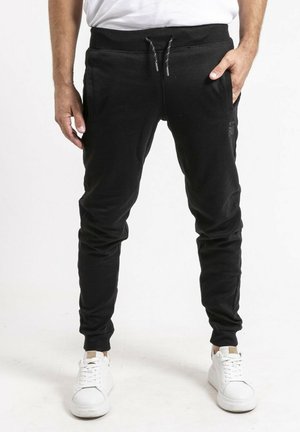 Homme portant un jogger noir à cordon de serrage et des baskets blanches, debout avec une main dans la poche.