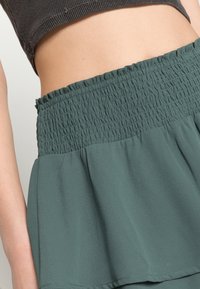 Una falda verde con una pretina fruncida y un diseño acampanado y plisado hecho de tela ligera. La textura es lisa y suave.