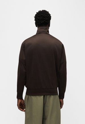Homme aux cheveux courts et bouclés, portant une veste marron foncé et un pantalon vert olive, debout, tourné de dos sur un fond uni.