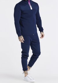 Tuta blu navy in tessuto morbido con giacca con zip e sezione del collo viola e bianca; i pantaloni hanno orli a costine e tasche.