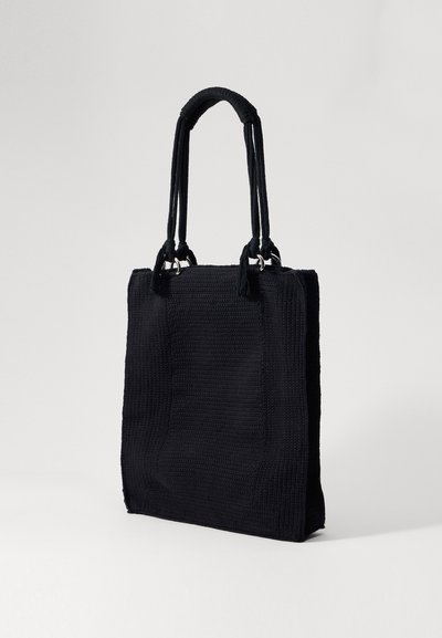 Black woven tote bag with textured surface, two sturdy handles, and decorative tassels at the base of the handles.