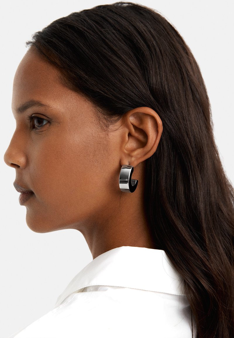 Czarna kolczyk w kształcie hoop, wykonany z polerowanego metalu, o gładkim, zaokrąglonym kształcie i subtelnym otwarciu. Noszony na uchu modelki.