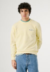 Gele sweatshirt met lange mouwen, ronde halslijn en een textuurstof. Bevat een klein geborduurd logo op de borst. Gecombineerd met blauwe jeans.