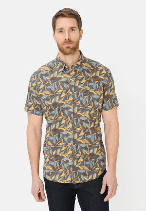 Mężczyzna ubrany w koszulę na guziki z krótkim rękawem z niebieskim, żółtym i brązowym wzorem liści, stojący na tle jasnego, jednolitego tła.