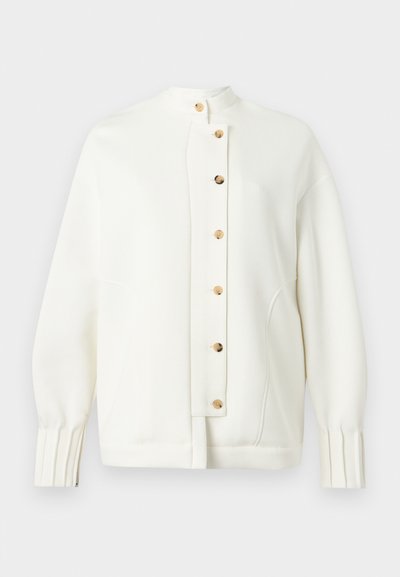 Λευκό μπουφάν από απαλό ύφασμα με ψηλό γιακά, με κούμπωμα με κουμπιά και χρυσά κουμπιά. Στρογγυλεμένους ώμους και πιέτες στα μανίκια.