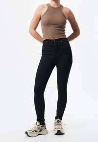 Svarta tajta jeans med hög midja, i kombination med en brun ribbad linne och beige sneakers med svarta detaljer och nätstruktur.