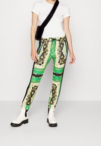 Pantalones estampados con un diseño barroco en verde, negro y dorado. El material parece ligero y con un ajuste relajado. Se llevan con una camiseta básica blanca y botas blancas.