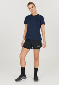 Marineblauw sportshirt gecombineerd met zwarte shorts met wit logo, zwarte crew-sokken en zwarte sportschoenen met textuurdetails.