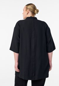 Schwarzes, übergroßes Hemd mit kurzen Ärmeln, das einen Kragen und ein gerades Rückendesign aufweist. Glattes Material mit lockerer Passform.