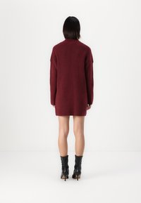 Bordeaux strikket kjole med høy krage, oversized design, ribbet tekstur og lange ermer, parret med svarte ankelstøvler.