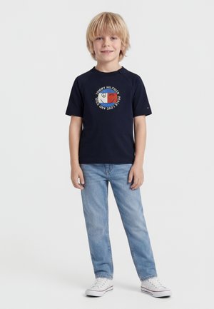 Ξανθός αγόρι που στέκεται φορώντας μαύρο t-shirt Tommy Hilfiger με σύμβολο της ειρήνης, ανοιχτό μπλε τζιν και λευκά αθλητικά παπούτσια μπροστά σε απλό φόντο.