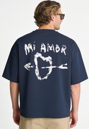 Mann mit welligem Haar und Sonnenbrille trägt ein marineblaues Hemd mit weißem Text "Mi Amor" und einem von einem Pfeil durchbohrten Herzen auf dem Rücken.