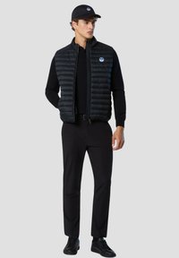 Chaleco acolchado negro con textura de acolchado vertical, llevado sobre una camiseta de manga larga negra. Combinado con pantalones y zapatos negros. Diseño simplista.