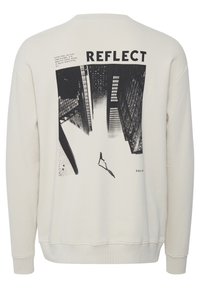 Sudadera de color beige claro con un diseño gráfico negro de edificios y una figura. La palabra "REFLECT" está impresa de manera destacada encima de la imagen.