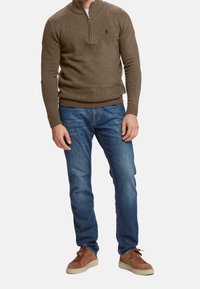 Uomo che indossa un maglione a metà zip marrone, jeans blu e sneaker in suede marroni, in piedi davanti a uno sfondo neutro.