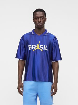 Tânăr purtând un tricou sport albastru cu "BRASIL" și un logo galben cu un jucător de baschet pe față, asortat cu pantaloni scurți bleu.