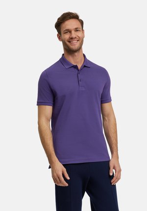 Polo shirt violet à manches courtes, avec un col et trois boutons au cou. Porté avec un pantalon bleu foncé. Texture lisse.