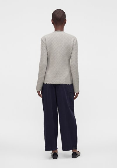 Personne debout de dos, portant un haut clair à manches longues texturé, un pantalon ample foncé et des chaussures noires, sur fond blanc uni.