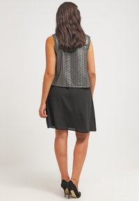 Černé bezrukávové šaty s volným střihem, vrstvené pod zkrácenou stříbrnou vestou s texturou. Černé boty na vysokém podpatku. Pohled zezadu.