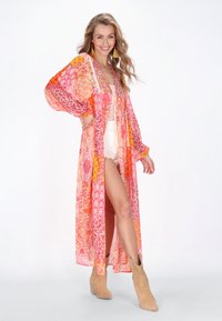 Kimono lungo e fluente nelle tonalità di rosa e arancione con motivi floreali. Tessuto leggero, apertura frontale e maniche ampie. Indossato con stivali alla caviglia color sabbia.