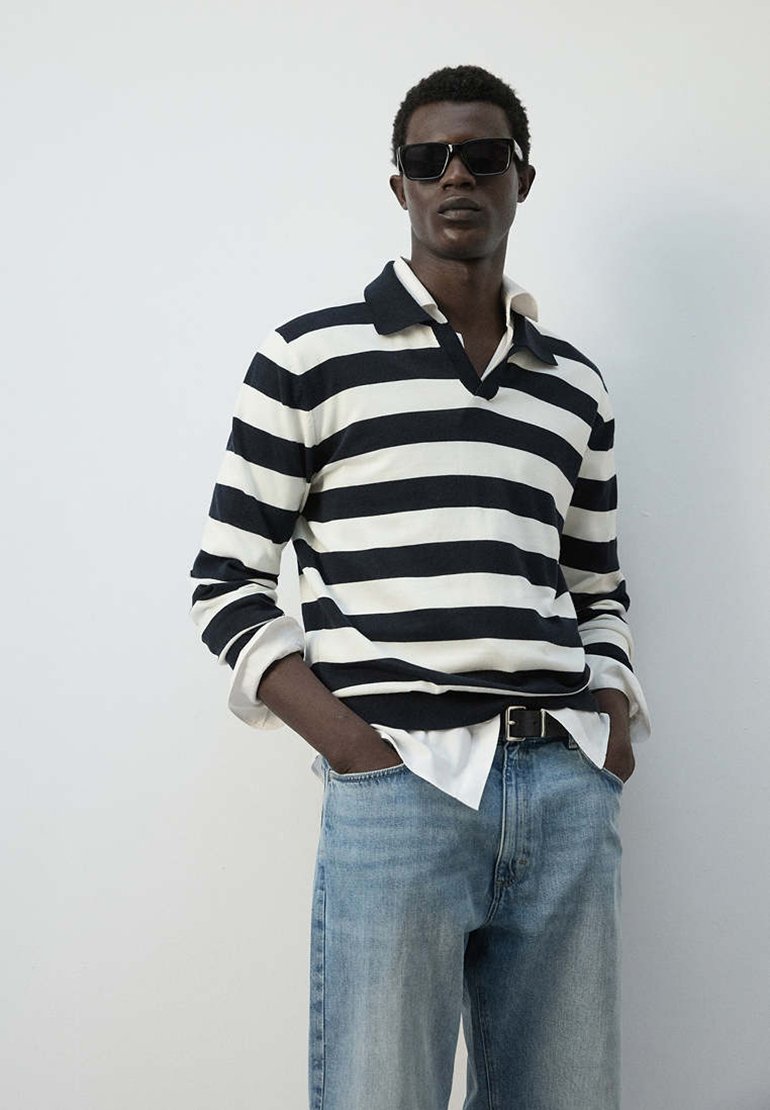 Homme portant des lunettes de soleil noires, une chemise à manches longues rayée noir et blanc par-dessus une chemise blanche, et un jean bleu clair avec les mains dans les poches.