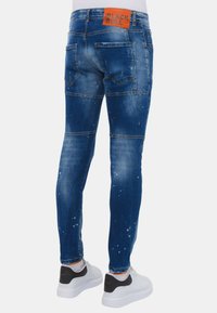 Skinny-fit jeans van middelblauwe denim met een vervaagde afwerking, versleten accenten en een oranje logo-opdruk op de taille. Gecombineerd met witte sneakers.