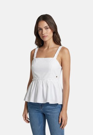 Giovane donna che indossa un top peplum bianco senza maniche con vita arricciata e jeans blu, in piedi davanti a uno sfondo chiaro e uniforme.