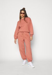 Roze katoenen sweatshirt met ballonmouwen, gecombineerd met joggers met trekkoord. Heeft elastische manchetten en witte sneakers met lichtblauwe accenten.