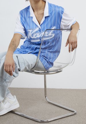 Chemise à rayures bleues et blanches avec un col boutonné, associée à un jean bleu clair déchiré et des baskets blanches, assise sur une chaise transparente.