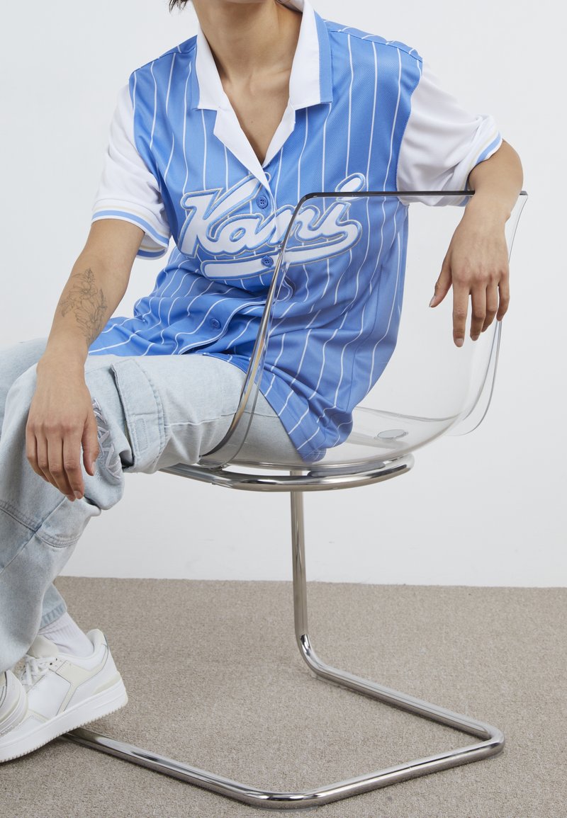 Chemise à rayures bleues et blanches avec un col boutonné, associée à un jean bleu clair déchiré et des baskets blanches, assise sur une chaise transparente.