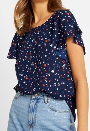 Femme portant un chemisier bleu marine à petites imprimés floraux rouges et blancs avec des manches évasées, rentrée dans un jean en denim délavé clair.