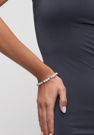 Woman’s hand resting on hip wearing a beaded bracelet with pale blue and gold beads, against a dark fitted garment background.