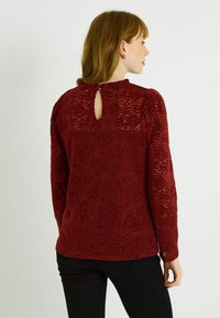 Haut à manches longues en dentelle rouge avec un col montant et un détail œillet dans le dos. Motif floral texturé sur toute la surface. Associé à un pantalon noir.