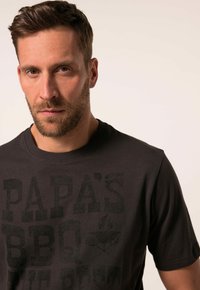 Dunkelgraues T-Shirt mit kurzen Ärmeln und Rundhalsausschnitt; zeigt eine markante schwarze Textgrafik mit der Aufschrift "PAPA'S BBQ THE BEST." Glatte Textur.