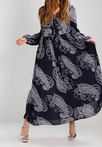 Dlouhé námořnické šaty s bílým paisley vzorem, které mají dlouhé rukávy a stažený pas. Stříbrné sandály na řemínky doplňují tento outfit.
