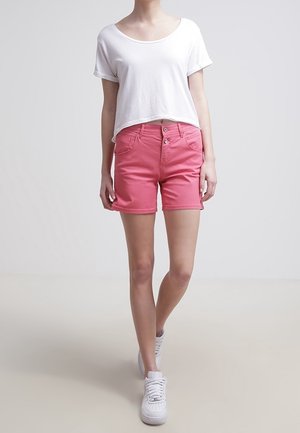 Shorts en denim rose clair avec une coupe décontractée, présentant un design à boutons et des poches avant, associés à un t-shirt blanc court et des baskets blanches.