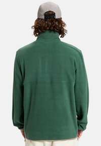 Pullover verde in fleece con colletto alto, maniche lunghe e superficie liscia. Presenta un delicato dettaglio delle cuciture sulla schiena.