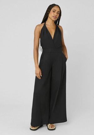 Femme aux longues tresses portant une combinaison sans manches noire à col halter avec jambes larges et sandales, debout avec une main dans la poche.