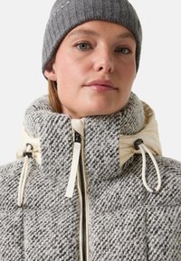 Jasno szara i kremowa kurtka w wzory z wysokim kołnierzem i regulowanymi sznurkami. Posiada przedni zamek błyskawiczny oraz szary czapkę z prążkowanego dzianiny.